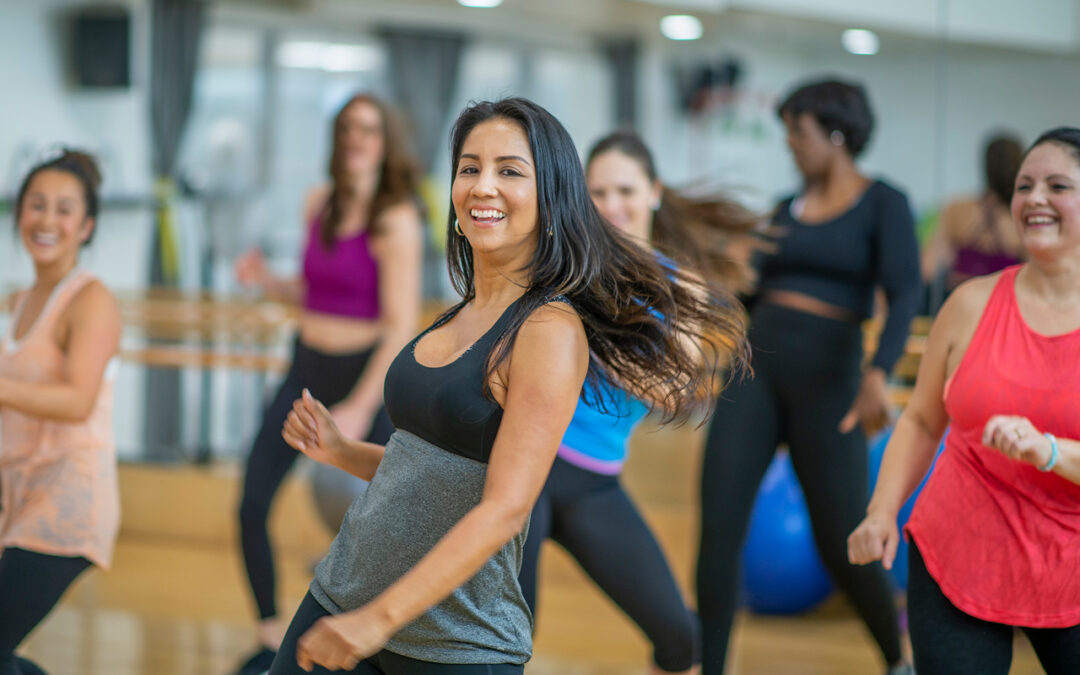 A multiethnic group of adult women are dancing in a fitness studio. They are wearing workout clothes. An ethnic woman is dancing in the foreground and smiling.